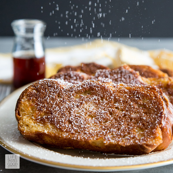 Oven French Toast on a white plate with a sprinkling of powdered sugar raining down. A bottle of syrup in the background