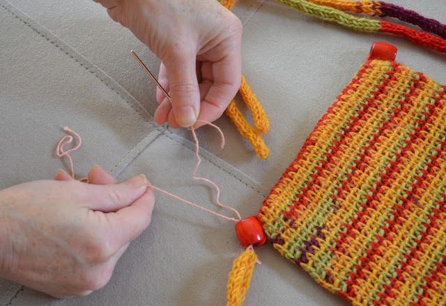 Two hands are used to thread the crocheted cord ends through the bead.  The right hand is holding the yarn tail while the left hand worked the needle and yarn through the bead, the drawstring ends and back into the beads.