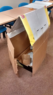 A cardboard anchorhold with an open doorway, revealing a bed and chamber pot, constructed from cardboard