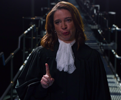 The Judge (Maya Rudolph), wearing a black judge's robes and a fluffy white collar thing, making a weird face where she's pulled her chin back so there are rolls in her neck, and she's holding up one finger of her right hand