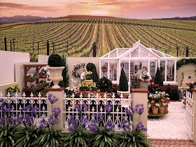 One-twelfth scale miniature garden courtyard with a greenhouse,a selection of plants and flowers and two men standing at the back. Behind the garden, vineyards stretch into the distance, towards the low sun. 