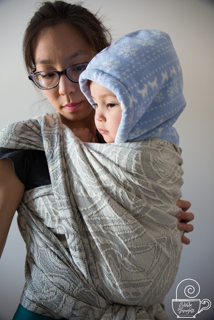 Image of a tan skin bespectacled Asian woman wearing a toddler on her front in a gray botanical-patterned woven wrap carrier with shoulder flips.