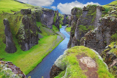 Paisaje de Islandia, donde un río cruza un valle de precipicios escarpados, con mucha hierba verde y un cielo estival.