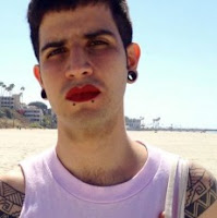 A young latino man with earplugs and red lipstick, standing on the beach in a pink tank top. Loma stands smiling lightly into the camera. 