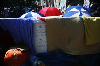 A photograph of a barrier with khaki blankets and blue bunting draped over it. A hand-lettered sign peeks out from behind the blankets. Only phrases on the sign are visible: [in French] "Come join... friends of... violently expelled..." Behind the barrier are red, white, and blue umbrellas and an indistinct crowd of people.