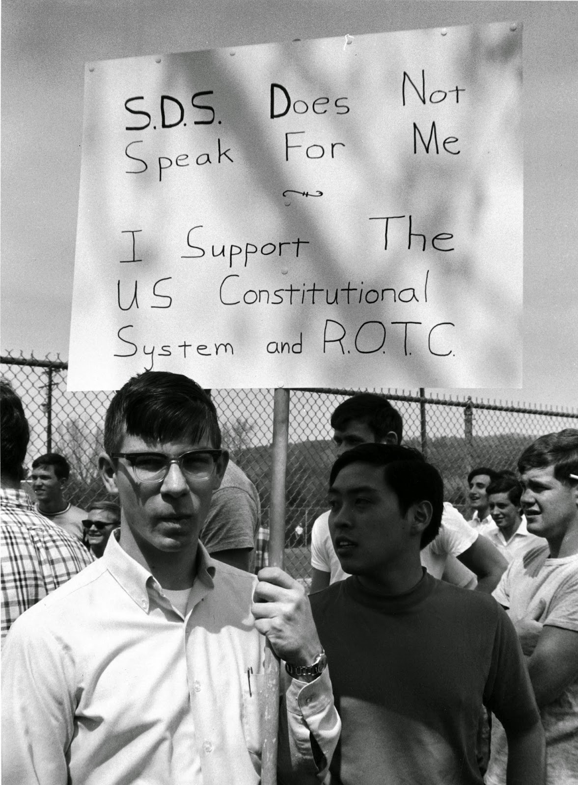 A photograph of a young man holding up a sign that reads "S.D.S. does not speak for me. I support the US Constitutional System and R.O.T.C."