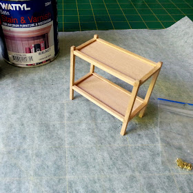 Modern miniature tea trolley on a workbench with a tin of stain and a bag of miniature castors next to it.