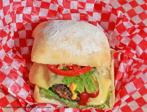 this is an Italian seasoned beef burger in a ciabatta bread roll pillow soft hamburger better than any fast food burger. This burger has lettuce tomato condiments, cheeses, and sitting on old fashioned white and red checkered wax paper
