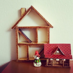 A miniature one-twelfth scale shadow box in the shape of a house displayed behind a tiny dolls house for a dolls house and a Toby dog figurine.