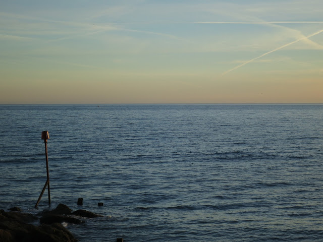 Looking out to sea in the evening with pink and yellow sky and a thing which looks like a standard lamp but isn't.