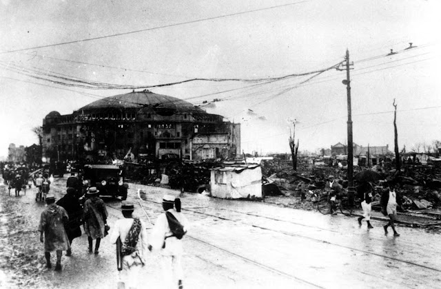 As pessoas andam na área devastada de Yuoguku, em Tóquio, Japão, após o terremoto que ocorreu em 1º de setembro de 1923. No fundo, está o edifício abobadado de cúpulas Kokugikan, arena de luta nacional de sumô, no distrito de Ryoguku.