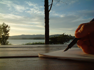 [scrivania di legno dove c'è biro e foglio ma anche un paesaggio di alberi,lago,cielo e colline]