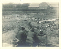 A black and white photograph of men from behind, looking over the edge of a trench.