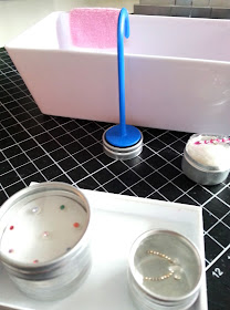 One-twelfth scale modern miniature bath with pink towel over the side and a floor-standing tap behind it, sitting on a small round tin lid. In the foreground is a small white tray containing a number of bindis in tiny tins.