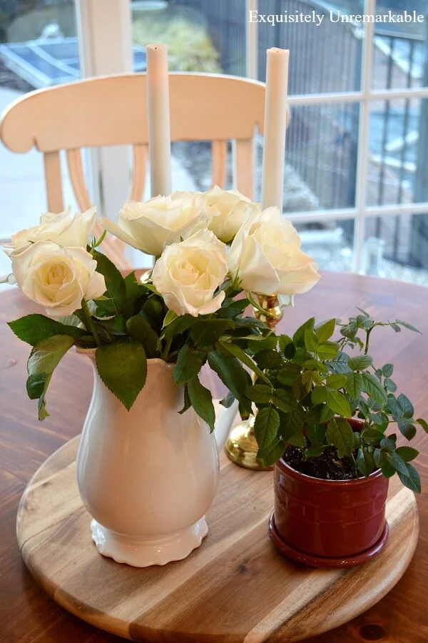 Mini rose bush in new pot on a lazy susan on table next roses in pitcher