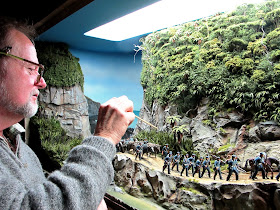 Man pointing a stick at miniature cabbage trees  in a diorama.