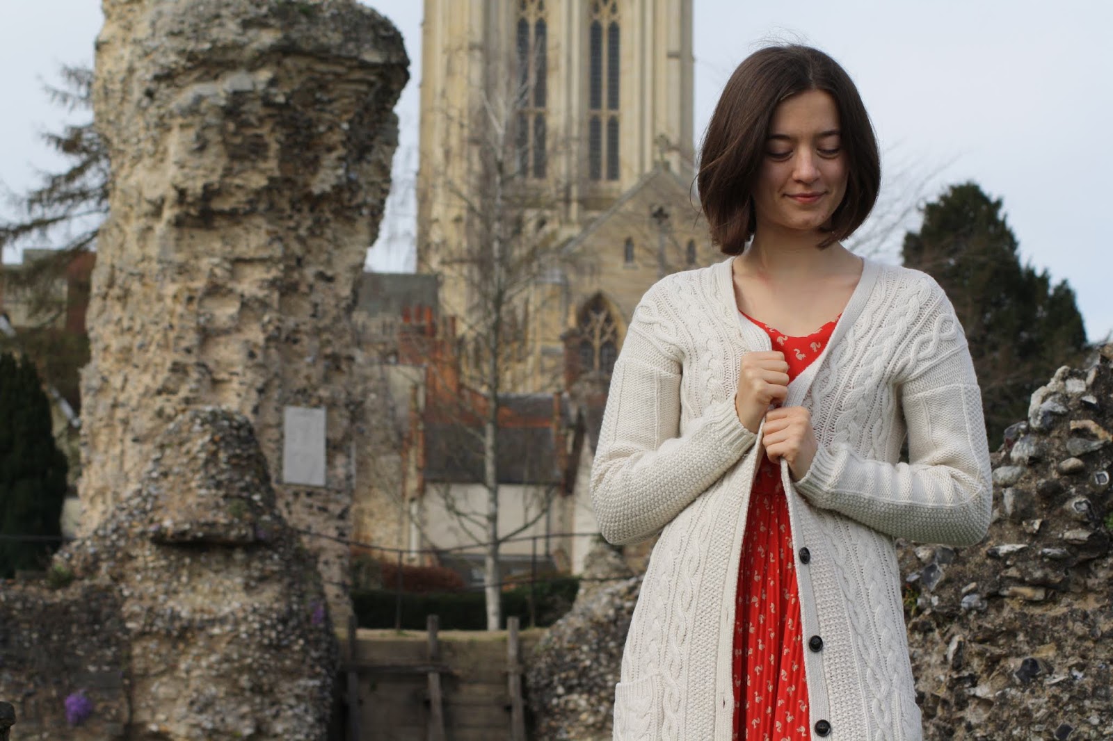 Abbey, wearing a red dress and knit cardi, stands in front of the ruins of Bury St Edmunds Abbey with the cathedral in the background