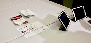 A pile of different rectangular A4 posters with different colours on each next to two bright white rectangular iPads on a bright white oval table on a bright background.