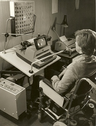 Possum Typewriter accessed using a sip and puff device and scan and select display. A young man in a wheelchair has a sip-puff tube in his mouth, looking at an illuminated alphabet and command board in front of him (via light-bulbs). A type-writer with magnifying glass sits between them.