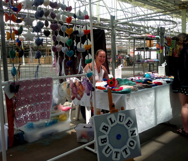 HandmadeByBlueButton stall with Kelly sitting behind a table smiling at the camera.  Items are displayed on the table and on racks either side. 