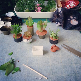 Five one-twelfth scale potted plants on a cutting mat surrounded by empty pots, spare foliage, a pack of air-drying clay and a cleaning rag.