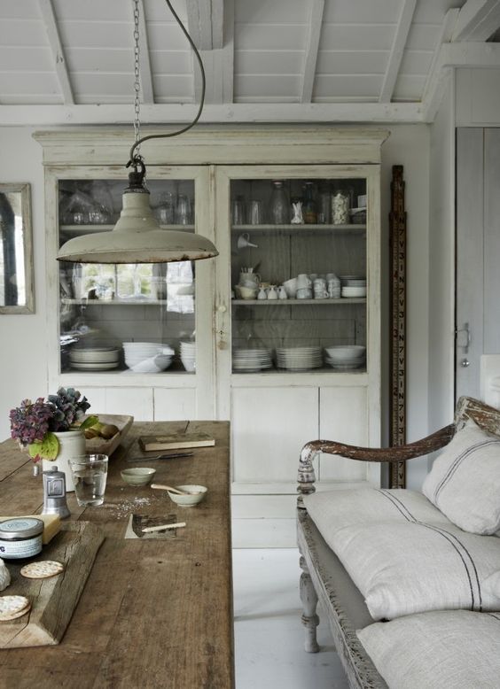 A gloriously rustic elegant kitchen with farm table, bench with linen grainsack pillows, and painted cupboard with dishes. A barn pendant light shimmers with vintage chic style. #frenchcountry #farmhousestyle #slowliving #countrydecor