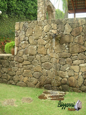 Tipo de muro de pedra com pedra bruta com a ducha d'água na bica de pedra com o piso de pedra em baixo da ducha com os pedregulhos do rio e o gramado de grama esmeralda.