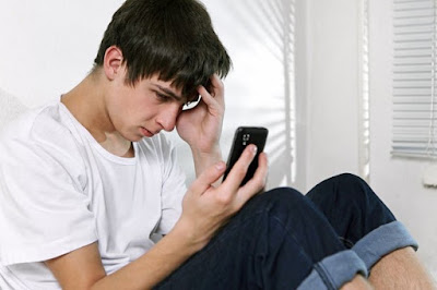 A boy with black hair, a white t-shirt, and black pants sits by his bedroom room looking anxiously at his phone.