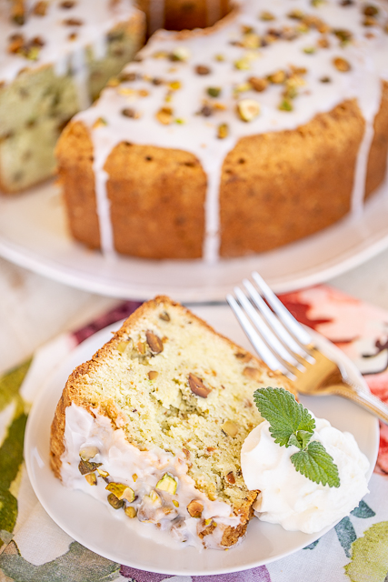 Pistachio Pound Cake - the most AMAZING pound cake I've ever eaten! So easy and delicious! Flour, eggs, sour cream, pistachio pudding, butter, sugar, vanilla, baking soda, salt, and chopped pistachios. Top the cake with a quick almond glaze for more amazing flavor. Serve the cake with whipped cream, mint and fresh berries. I took this to a party and everyone asked for the recipe! #poundcake #dessert #pistachio #potluck slice of pound cake on a plate with cake in the background