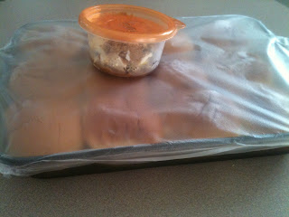 A baking sheet with ham and cheese sandwiches covered with plastic wrap, with a small container of seasoning sitting gone top.
