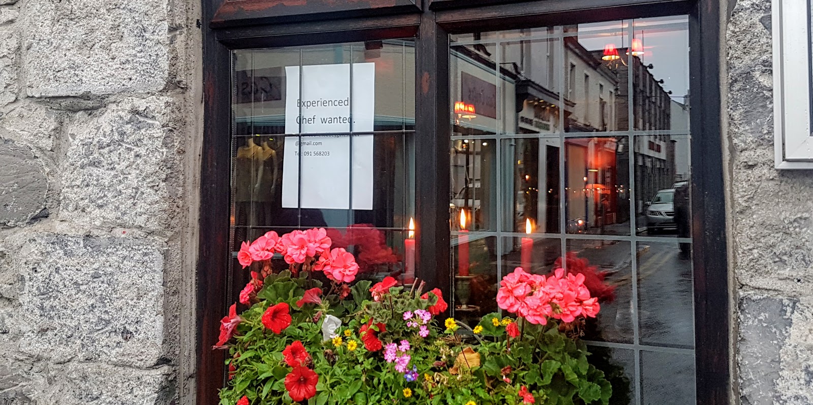 restaurant window with pink flowers in the window box, candles inside - and a chef-wanted sign