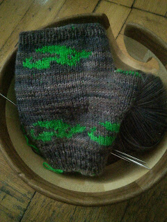 A fingerless glove knit in brown yarn, with a small dragon motif done in green yarn.  The stitches for the thumb of the glove are on scrap yarn.  The yarn balls and glove are set inside a wooden yarn bowl.