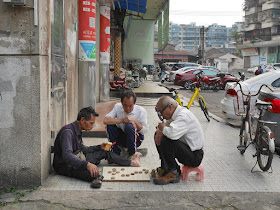 man holding cigarettes and a man with a tobacco pipe playing a game of xiangqi