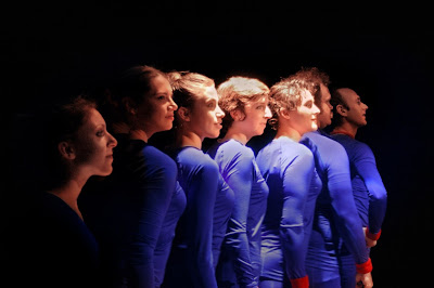 Seven people in purple-blue leotards lined up against a black background