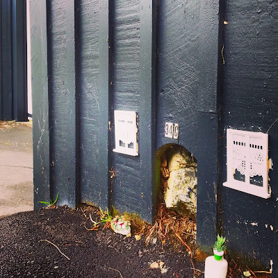 Wooden fence with a hole cut in it to accommodate a jutting piece of concrete, and the numbers '34 C' added above. Next to the hole are glued two dolls' house windows with blinds.