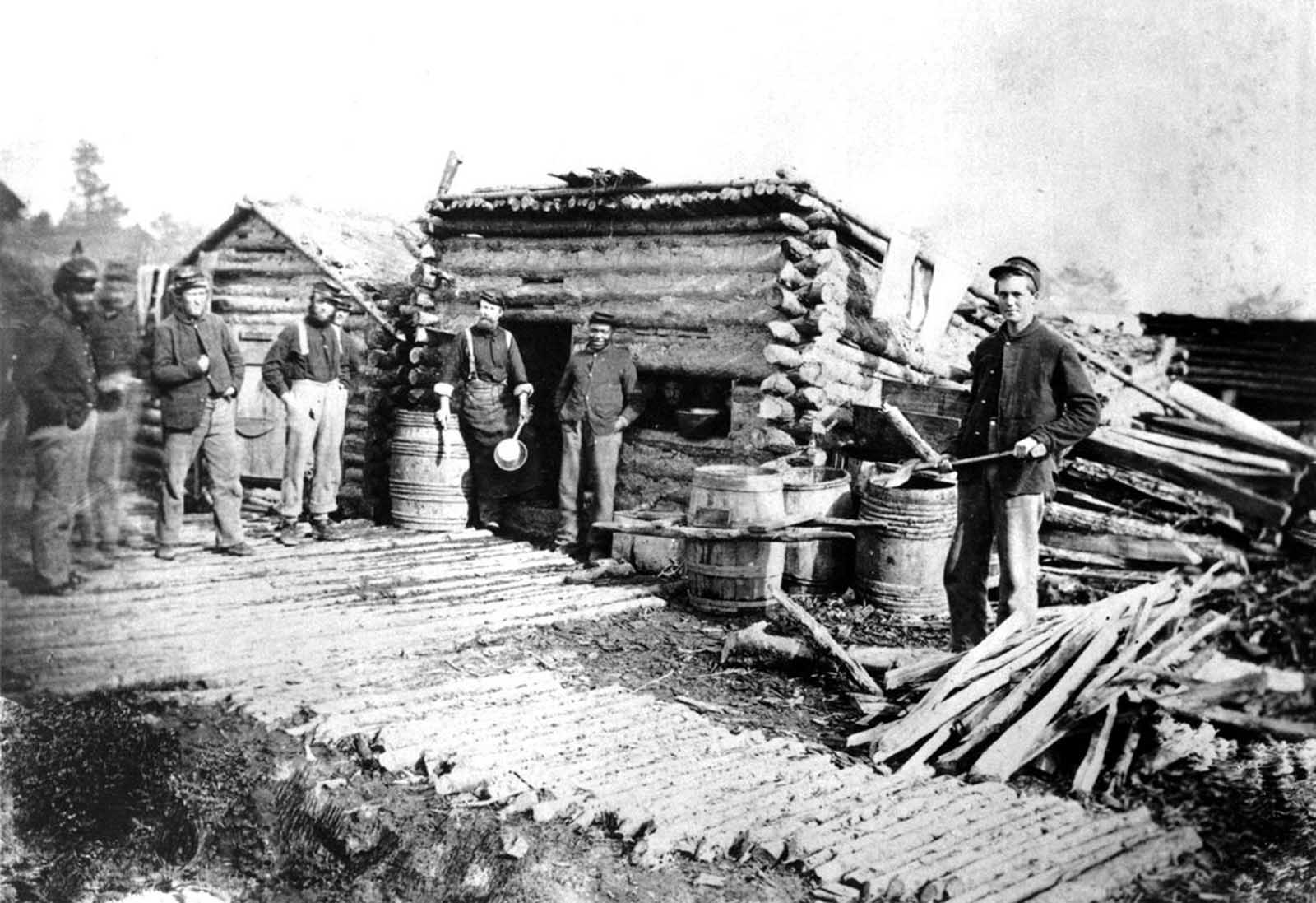 Serving as a soldier in uniform and getting regular army pay, a former slave (center, with hands in pockets) stands with other Federal soldiers at the Army of the Potomac winter headquarters near Fredericksburg, Virginia, The log hut served as a mess house for the regiment.