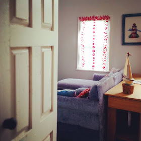 View through the door of a one-twelfth scale modern miniature house, into the lounge where you can see a grey velvet sofa underneath a window with a red and white net curtain at it. Behind the sofa is a wooden sideboard with a model sailboat on it. And above it is a corss stitch picture of a lighthouse.