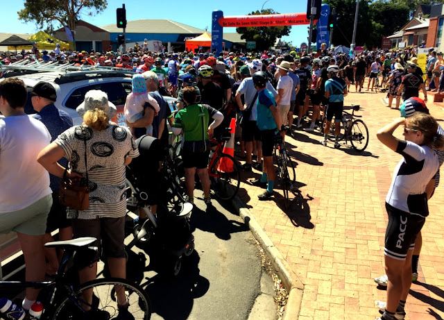 The blue and red starting line banner is surrounded by spectators on both sides of the footpath. At certain points close to the starting line, crowds are around 10 people deep, maybe more. The picture shows a sea of riders and crowds.