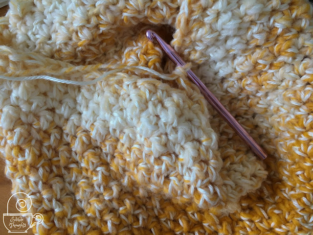 Image of a crocheted work in progress that's yellow and cream attached to a pink metal crochet hook.