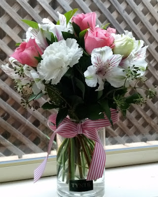 A pretty bunch of pink and white flowers in a plain glass vase decorated with a pink and white candy-striped ribbon. The vase is sitting on the windowsill with a trellis outside the window in the background. The flowers are white and pink lilies, white carnations and pink roses with green filler (maybe gyspophila - the flowers are still buds). The florist's name is printed on a label and stuck onto the bottom front of the jar.