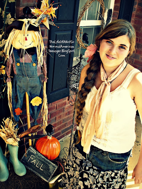 Photo of a teen girl with long brown hair braided over one shoulder and down past her waist. She has a scarf around her neck and has a white shirt on and a blue and white skirt. She is standing on a porch with autumn decor one it. There is a scarecrow, an orange pumpkin, a sign that says "fall". A pair of light blue rain boots are near and there are dried corn in them. The article is Thanksgiving Recipes by RosevineCottageGirls.com