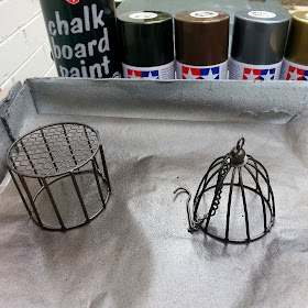 Two painted wire items on a sheet of baking paper in a box, with a row of various spray cans lined up behind in browns, greys and metal colours.