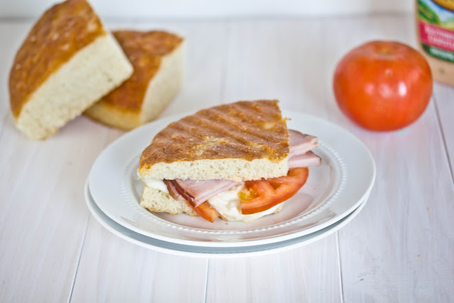 A sandwich on a plate with bread and tomato in the background