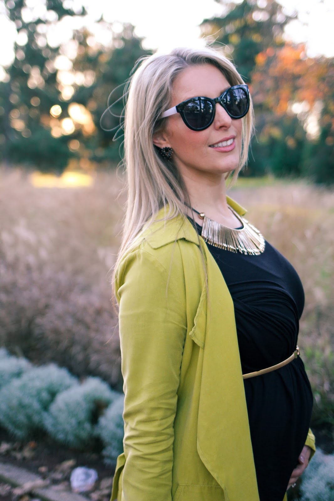 The Goldfields Girl styling maternity fashion. She wears black maternity dress, green jacket, statement gold necklace, brown boots and Michael Kors bag.