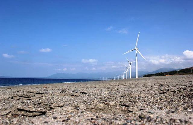 Windmill align and standing tall beside the shore and a view of distant mountains under the clear blue sky.