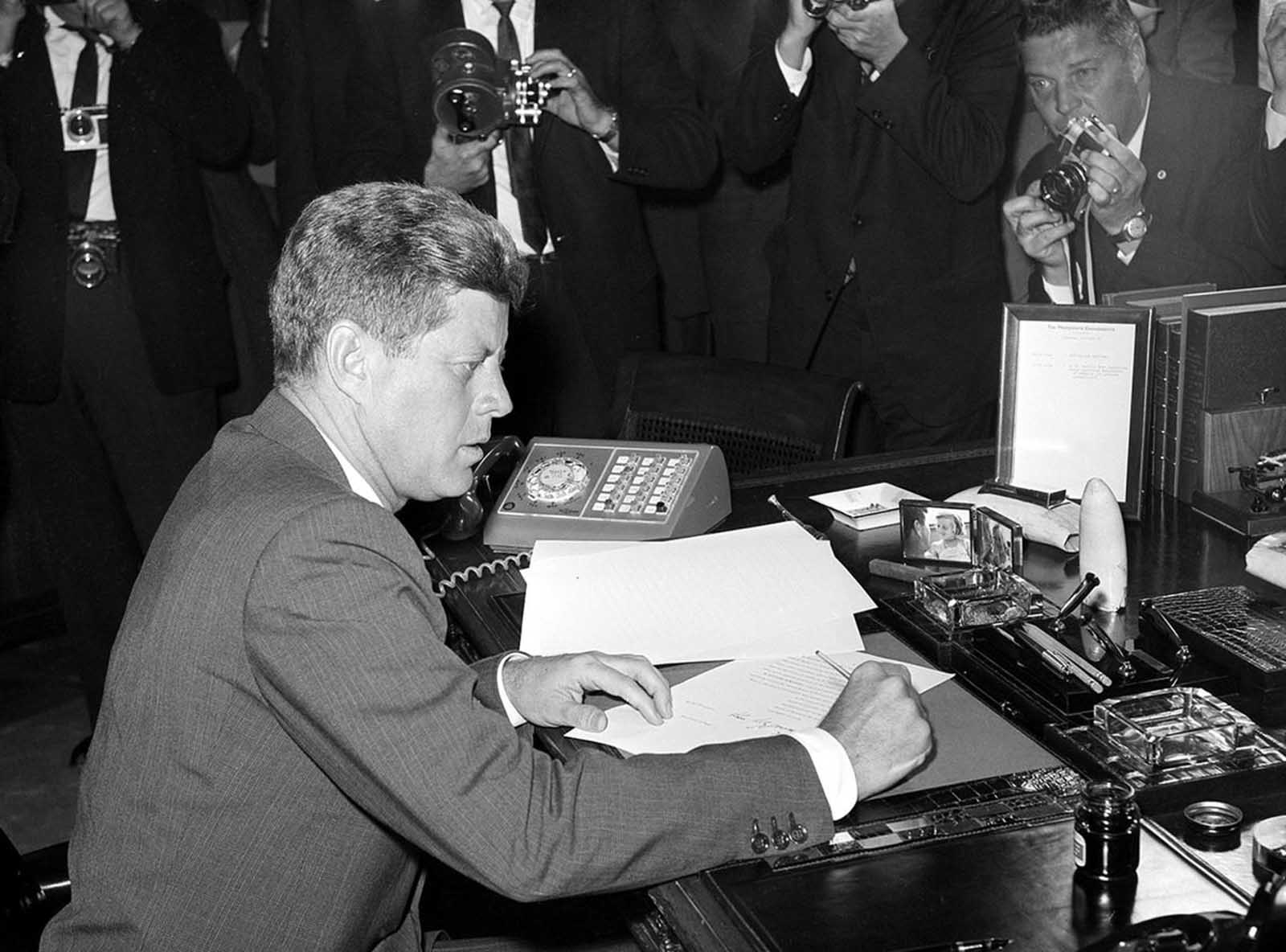 President Kennedy is surrounded by photographers as he sits at his desk in the White House, on October 23, 1962, shortly after signing a presidential proclamation concerning the Cuba Missile crisis.