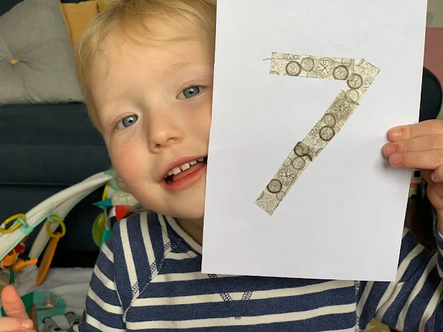 Toddler holding up a sheet of paper with the number 7 covered in washi tape