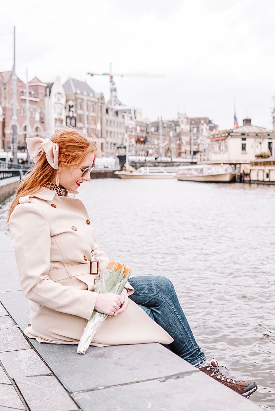 tampa blogger amanda burrows is in amsterdam. she is sitting along the canal. she is wearing a tan trench coat and holding orange tulips.