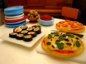 Miniature dolls' house savoury food arranged on a table, including sushi, nuts, olives and two sorts of pizza.