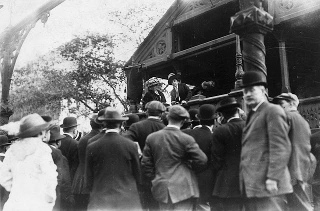 Mrs. E.R. Smith practicing speechmaking from a covered platform before a small crowd, a 'school for suffragette speakers' in Union Square.
